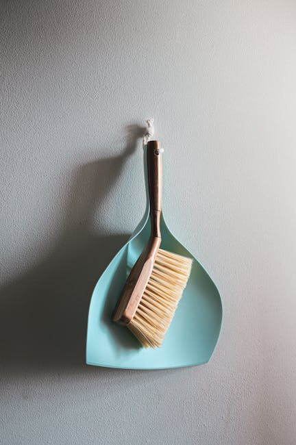 A close-up of a light blue dustpan and a wooden-handled brush hanging on a white wall, used for surface cleaning and debris collection. The dustpan appears clean and dry, with a smooth surface, while