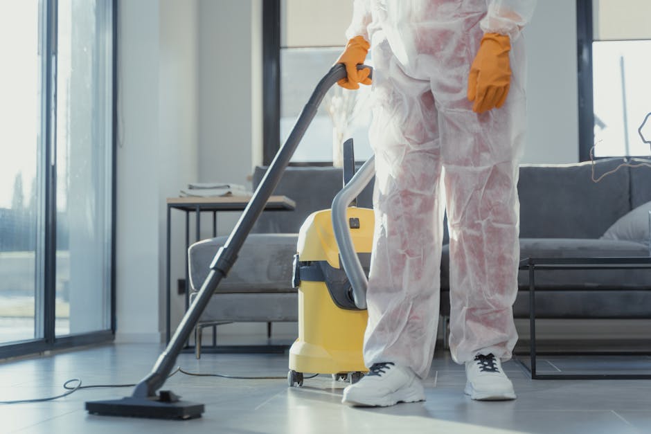 A person wearing bright yellow rubber gloves holding a clear glass spray bottle with a red nozzle and a white trigger, along with a blue cleaning sponge, positioned over a plain light gray surface. Th