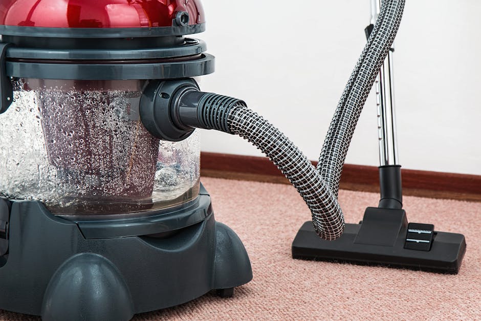 A person wearing light-colored shoes and denim jeans is using a handheld vacuum cleaner to clean an ornate, patterned carpet in a room. The carpet features a floral design in muted tones of beige, gre